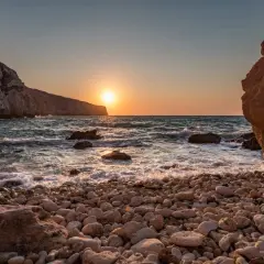 Malta zachód słońca nad kamienistą plażą. Słońce odbija się w falach, a klify tworzą malowniczą scenerię.