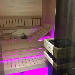 Drewniana sauna z różowym podświetleniem. Widoczny piec, ławki i wiaderko. Czy wolisz saunę suchą czy mokrą?