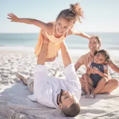 Rodzina na plaży na Malcie. Ojciec podrzuca córkę, matka z drugą córką siedzą obok. Czyste niebo, spokojne morze, piasek. Malta bezpieczeństwo.