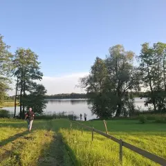 Piękny litewski krajobraz nad jeziorem. Osoba idzie ścieżką przez zieloną łąkę w stronę wody, gdzie stoją inni ludzie.