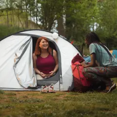 Dwie kobiety rozmawiają przy namiocie, planując, gdzie pod namiot z dziećmi w Polsce. W tle inne namioty i las.