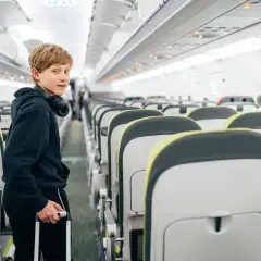 Chłopiec z słuchawkami na szyi i walizką w ręku stoi w samolocie. Zastanawia się, do jakiego wieku dzieci latają za darmo.