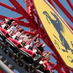 Ludzie krzyczą z radości na czerwonym wagoniku, pędzącym po torach najwyższego rollercoastera w Europie, obok żółtego logo Ferrari.
