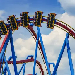 Wagoniki największego rollercoastera na świecie pędzą po czerwono-żółtych torach na tle błękitnego nieba z chmurami.