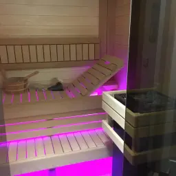 Drewniana sauna z różowym podświetleniem. Widoczny piec, ławki i wiaderko. Czy wolisz saunę suchą czy mokrą?