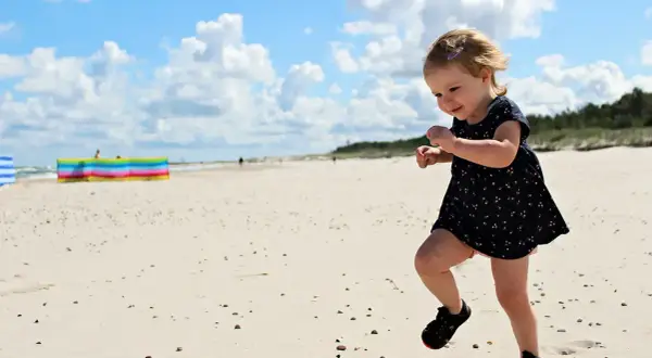 Radosna wycieczka dla dzieci na plażę. Mała dziewczynka w letniej sukience biega po piasku, ciesząc się słońcem i chmurami.