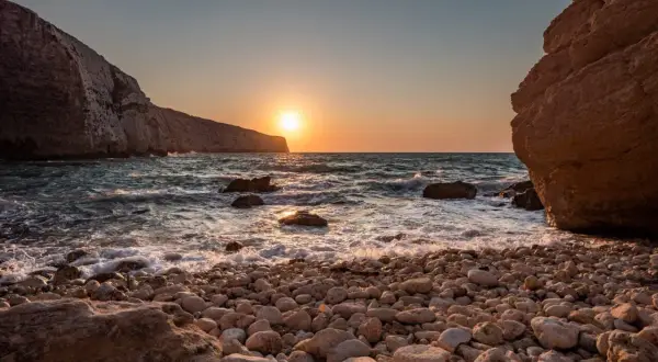 Malta zachód słońca nad kamienistą plażą. Słońce odbija się w falach, a klify tworzą malowniczą scenerię.