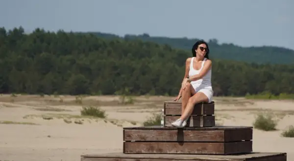 Kobieta w białej sukience siedzi na drewnianych schodkach, podziwiając krajobraz przypominający polską sahara.