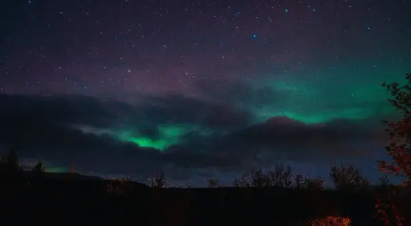 Zielona zorza polarna tańczy na nocnym niebie nad Islandią, wśród gwiazd i ciemnych chmur.
