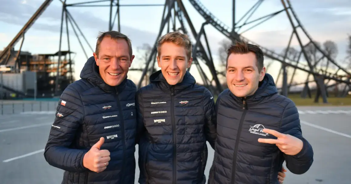 Trzech mężczyzn w kurtkach z logo Energylandii pozuje na tle konstrukcji roller coastera.