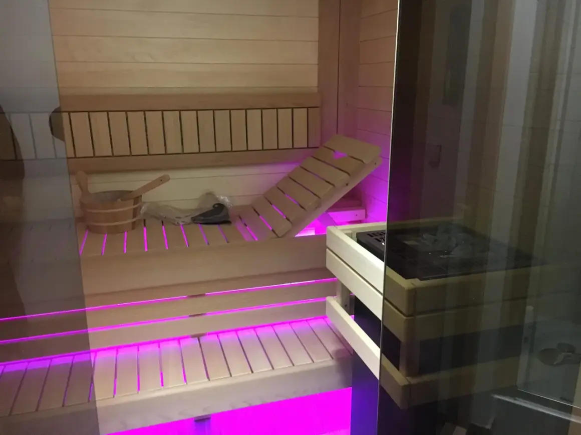 Drewniana sauna z różowym podświetleniem. Widoczny piec, ławki i wiaderko. Czy wolisz saunę suchą czy mokrą?