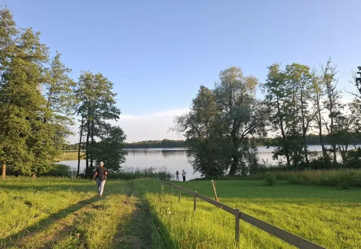 Piękny litewski krajobraz nad jeziorem. Osoba idzie ścieżką przez zieloną łąkę w stronę wody, gdzie stoją inni ludzie.