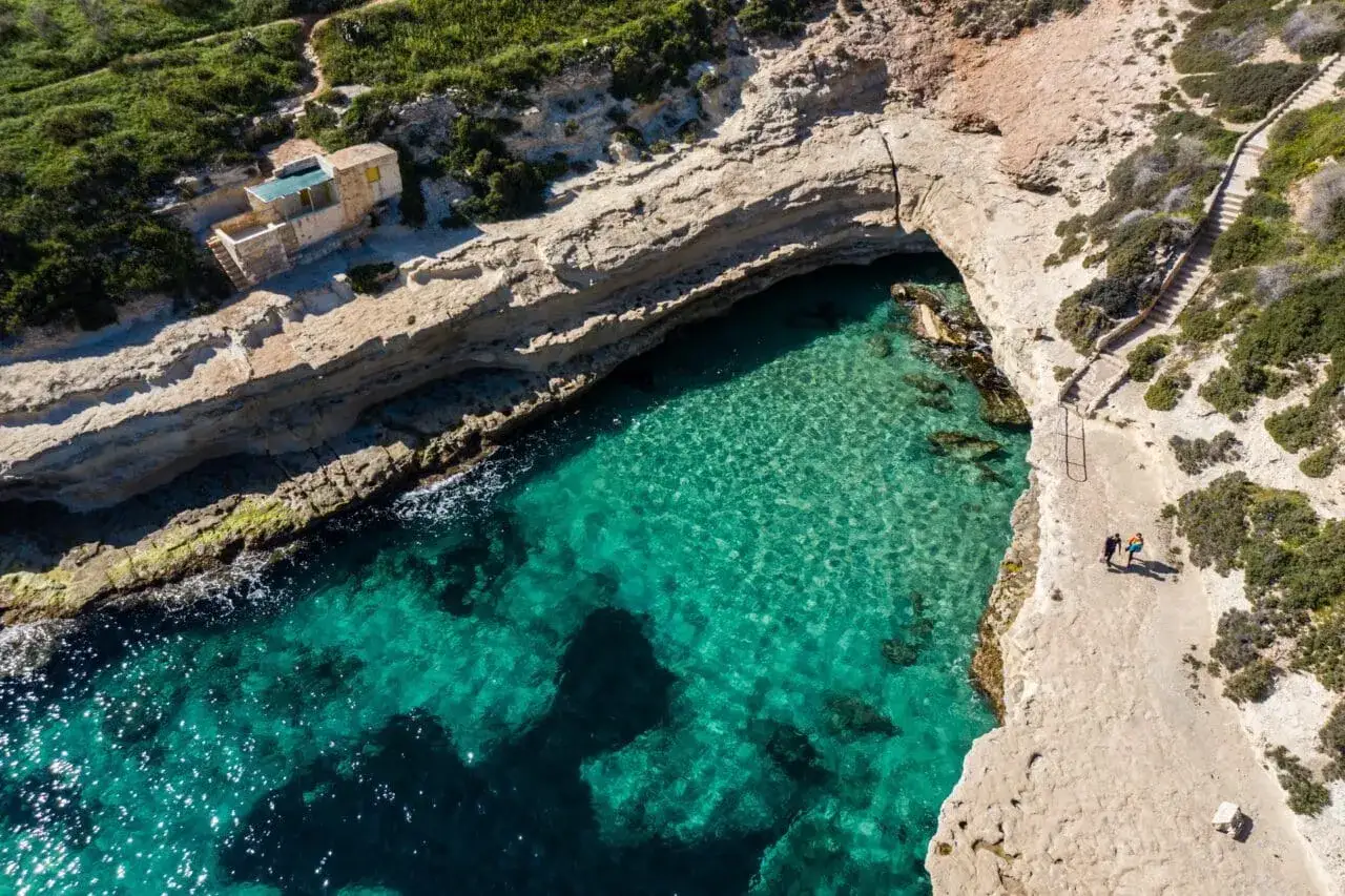 Malta: turkusowa zatoka, skaliste wybrzeże i schody prowadzące do wody. Dwie osoby spacerują ścieżką, czując bezpieczeństwo i spokój.