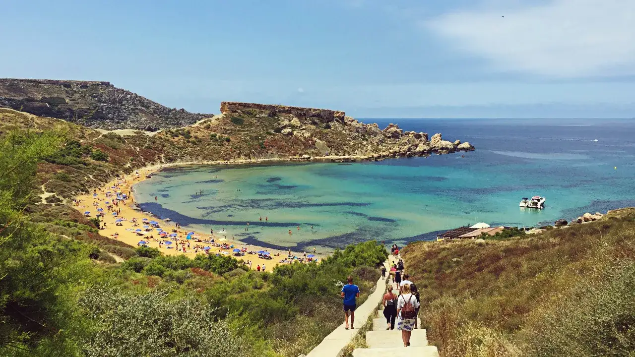 Malta: malownicza zatoka z turkusową wodą, piaszczystą plażą i ludźmi schodzącymi po schodach. Czy na Malcie jest drogo? Warto sprawdzić ceny przed wyjazdem.