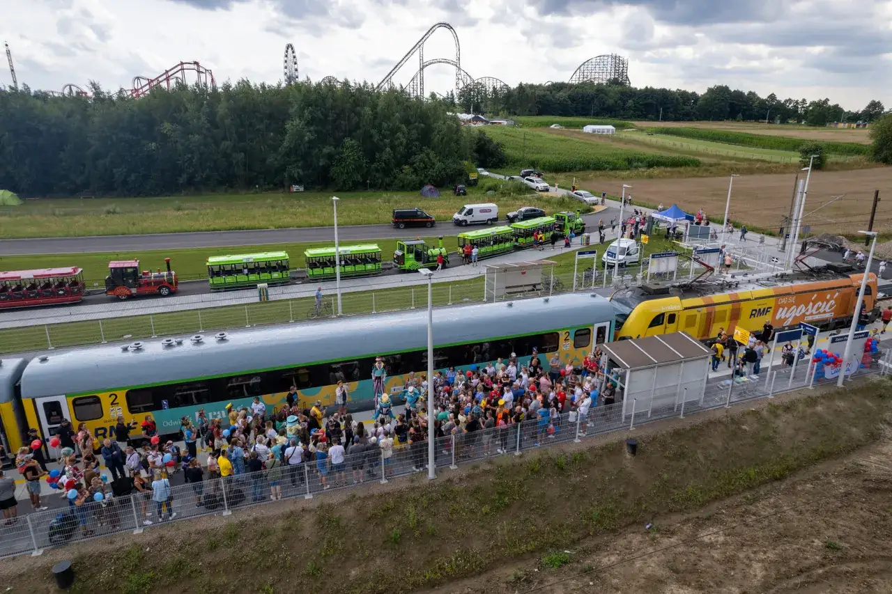 Pociąg do Energylandii przyjechał, a tłum podekscytowanych pasażerów czeka na wejście. W tle widać kolejki górskie.