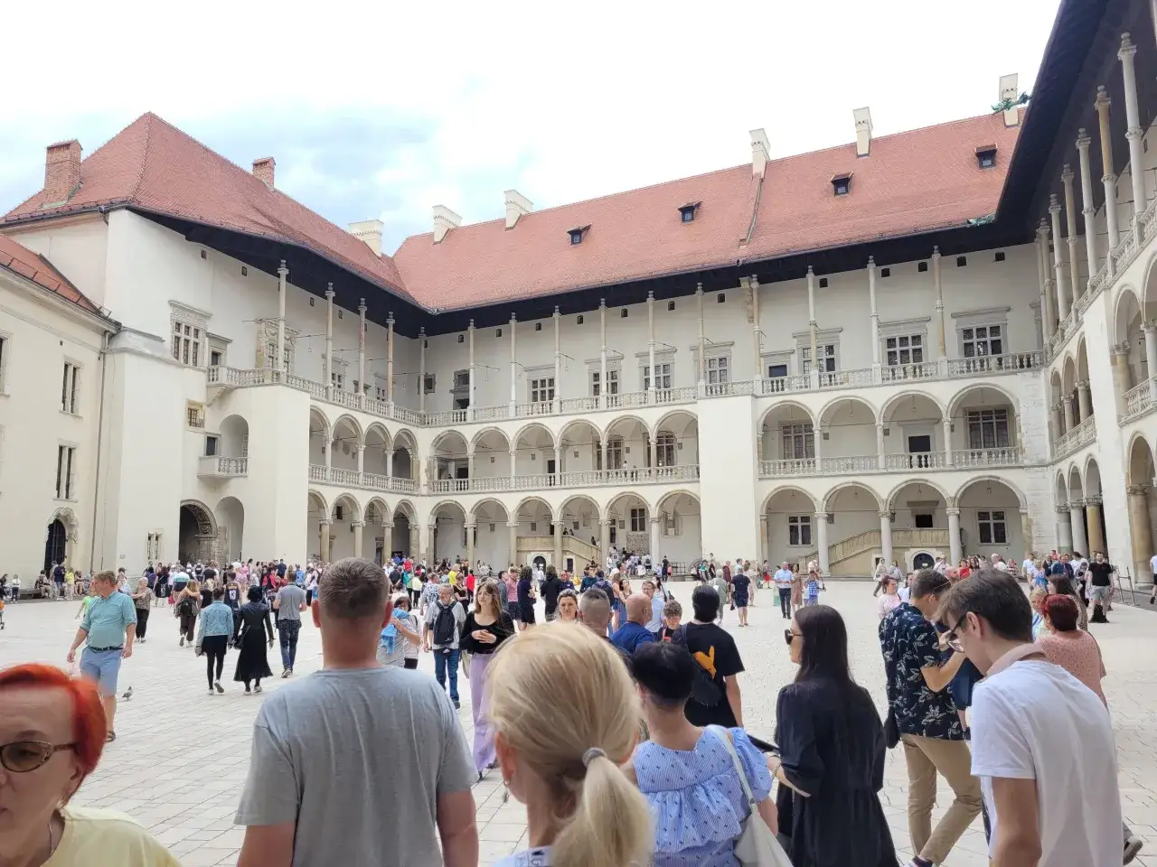 Tłum turystów zwiedza dziedziniec Wawelu. Grupa słucha opowieści przewodnika, podziwiając renesansową architekturę.