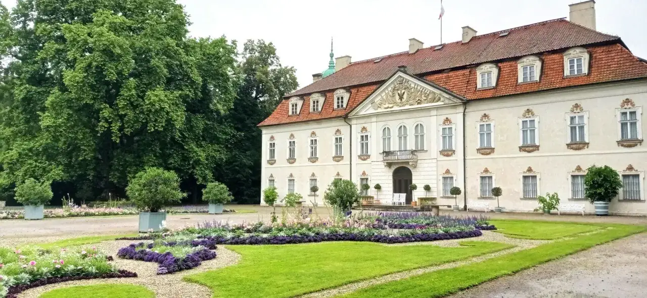 Pałac w Nieborowie otoczony kwitnącym ogrodem z klombami pełnymi fioletowych i różowych kwiatów. W tle wysokie drzewa.