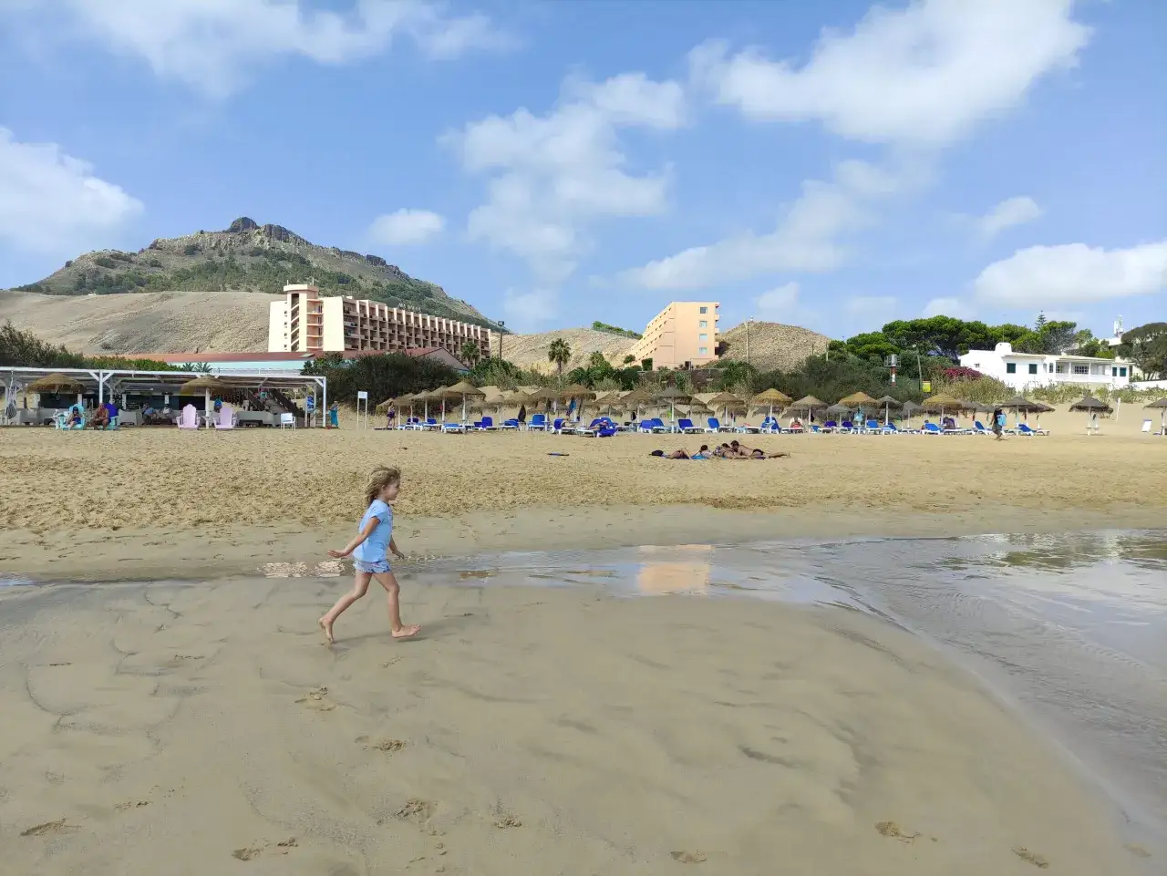 Dziewczynka biega po plaży, w tle hotele i góry. Madera z dzieckiem w słoneczny dzień.