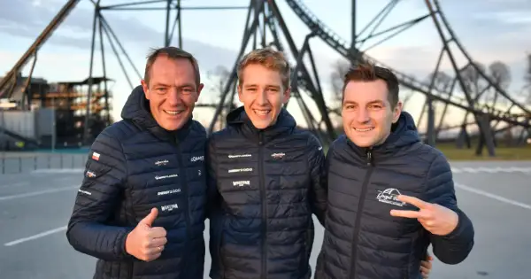 Trzech mężczyzn w kurtkach z logo Energylandii pozuje na tle konstrukcji roller coastera.